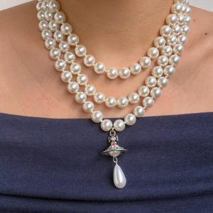 Op de afbeelding: Een parelketting met drie strengen bestaande uit ronde witte kralen. Een zilverkleurige orb-hanger met kleurrijke accenten en een bungelende druppelvormige parelbedel hangt aan het midden van de onderste streng, rustend op het sleutelbeen.