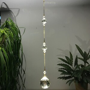 May include: A clear crystal suncatcher hanging from the ceiling. The suncatcher is made of multiple clear crystal beads and orbs strung together with a gold-colored chain. The bottom orb is a large, faceted crystal ball.