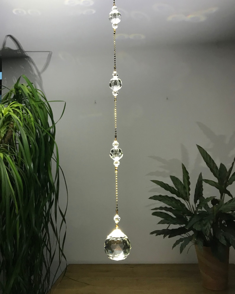 May include: A clear crystal suncatcher hanging from the ceiling. The suncatcher is made of multiple clear crystal beads and orbs strung together with a gold-colored chain. The bottom orb is a large, faceted crystal ball.