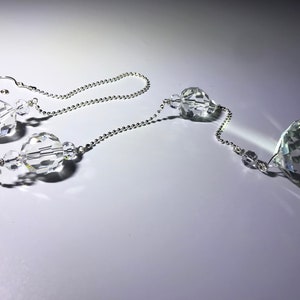May include: Clear crystal suncatcher with a faceted, teardrop-shaped pendant and multiple crystal beads strung on a silver-colored chain. The suncatcher is designed to hang and catch the light.