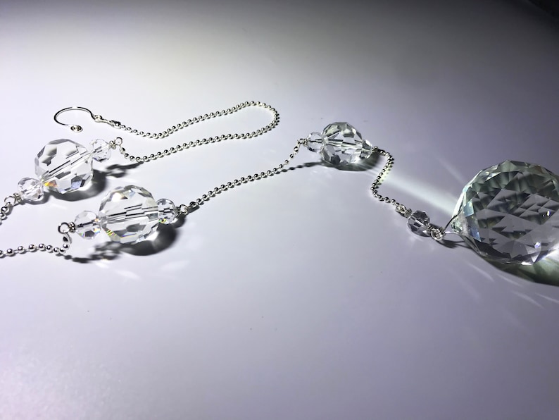 May include: Clear crystal suncatcher with a faceted, teardrop-shaped pendant and multiple crystal beads strung on a silver-colored chain. The suncatcher is designed to hang and catch the light.