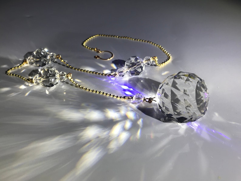 May include: A clear crystal ball suncatcher with a faceted design, suspended by a gold-colored chain. Additional clear crystal beads are strung along the chain. The suncatcher casts light patterns on the surface.