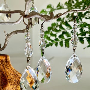 Puede incluir: Pendientes de araña de cristal transparente con colgantes facetados en forma de lágrima y cuentas más pequeñas. Los pendientes cuelgan de ganchos de color bronce y se exhiben en una rama decorativa con un fondo borroso de vegetación.