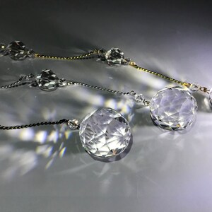 May include: Close-up of three strands of crystal ball garlands. Each strand features faceted crystal spheres of varying sizes, strung together with delicate chains in gold, silver, and black. The crystals reflect light, creating a sparkling effect.