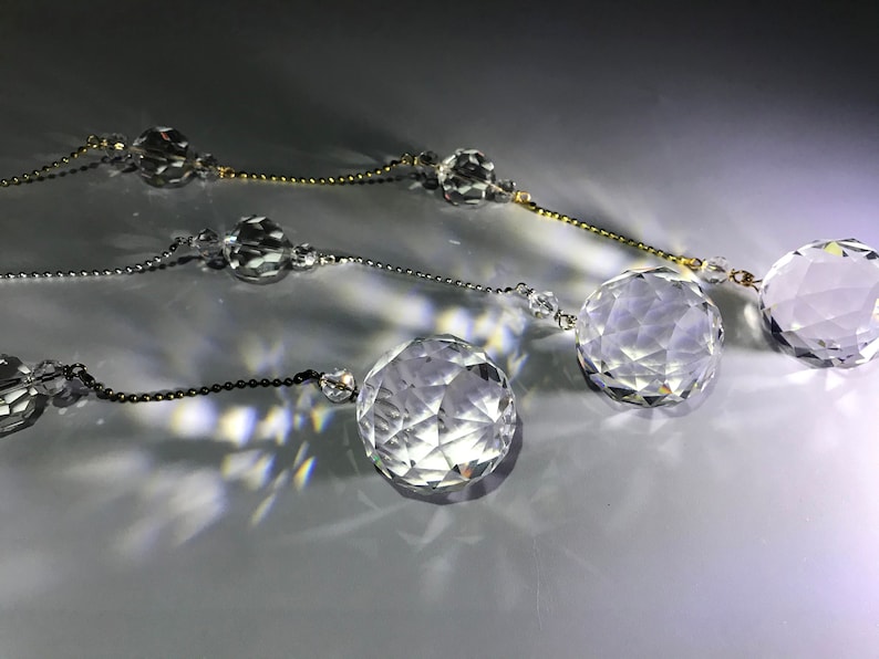 May include: Close-up of three strands of crystal ball garlands. Each strand features faceted crystal spheres of varying sizes, strung together with delicate chains in gold, silver, and black. The crystals reflect light, creating a sparkling effect.
