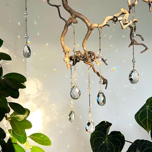 Könnte beinhalten: Fünf kristallene Tropfen-Anhänger hängen an einem Ast, jeder an einer zarten goldenen Kette. Die Kristalle fangen das Licht ein und erzeugen einen schimmernden Effekt.