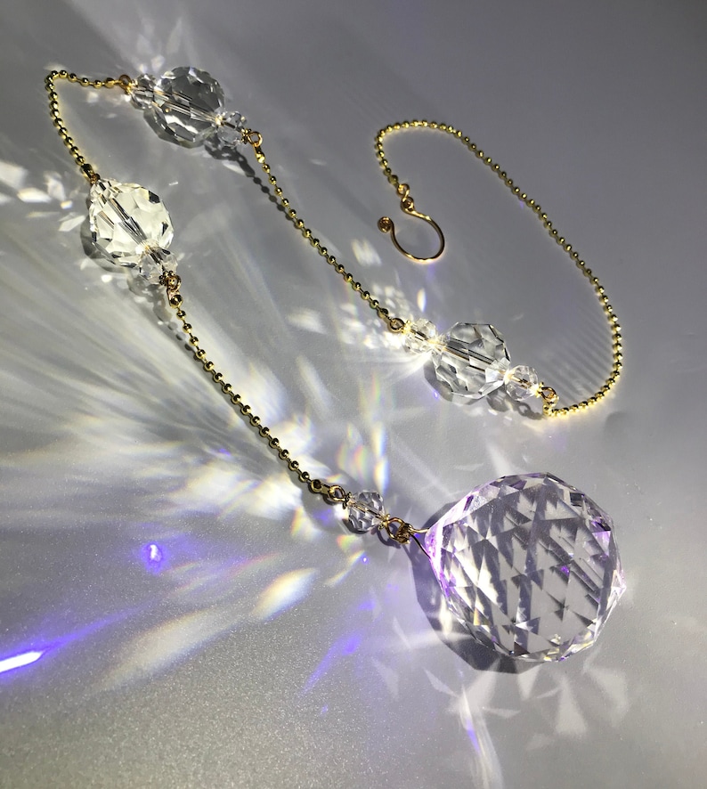 May include: A clear crystal suncatcher with a faceted sphere at the bottom, connected by a gold chain to other crystal beads and a hook. The suncatcher is casting light reflections on a silver surface.
