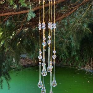 K&ouml;nnte beinhalten: Ein Kristall-Tropfen-Kronleuchter mit mehreren Str&auml;ngen goldener Kette und klaren Kristallperlen. Der Kronleuchter h&auml;ngt an einem Ast &uuml;ber einem gr&uuml;nen Teich.