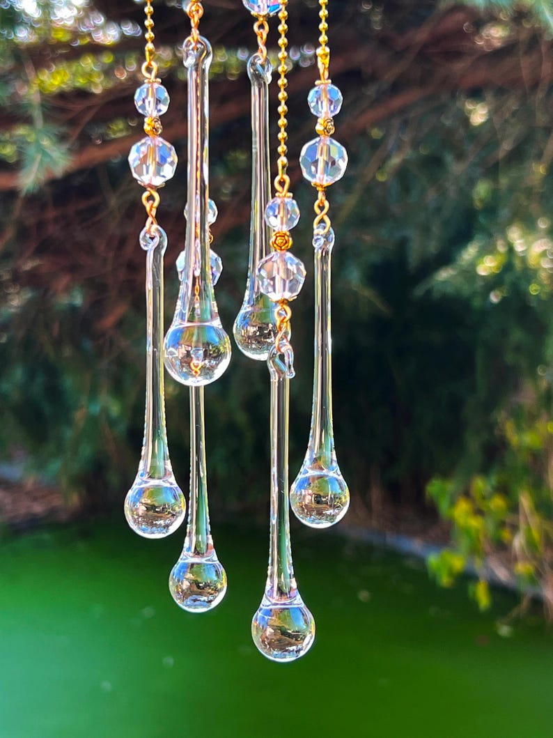 K&ouml;nnte beinhalten: Ein Kristall-Sonnenf&auml;nger mit mehreren tropfenf&ouml;rmigen Prismen, die an einer goldenen Kette h&auml;ngen. Die Prismen sind klar und reflektieren das Licht.