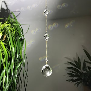 Puede incluir: Un atrapasoles de cristal transparente con una gran esfera facetada en la parte inferior, conectada por una cadena dorada a pequeñas cuentas de cristal. El atrapasoles cuelga frente a una pared blanca, con una planta verde al fondo.