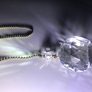 May include: A clear, faceted crystal ball ornament hangs from a delicate gold-colored chain. The crystal sphere reflects light, creating a sparkling effect. The background is a gradient of dark and light tones.