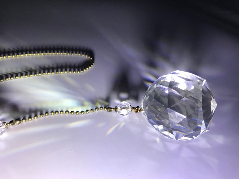May include: A clear, faceted crystal ball ornament hangs from a delicate gold-colored chain. The crystal sphere reflects light, creating a sparkling effect. The background is a gradient of dark and light tones.