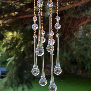 K&ouml;nnte beinhalten: Ein Kristall-Tropfen-Sonnenf&auml;nger mit f&uuml;nf Str&auml;ngen goldener Kette und klaren Kristallperlen. Der Sonnenf&auml;nger h&auml;ngt an einem Baumzweig und der Hintergrund ist ein gr&uuml;ner Teich.
