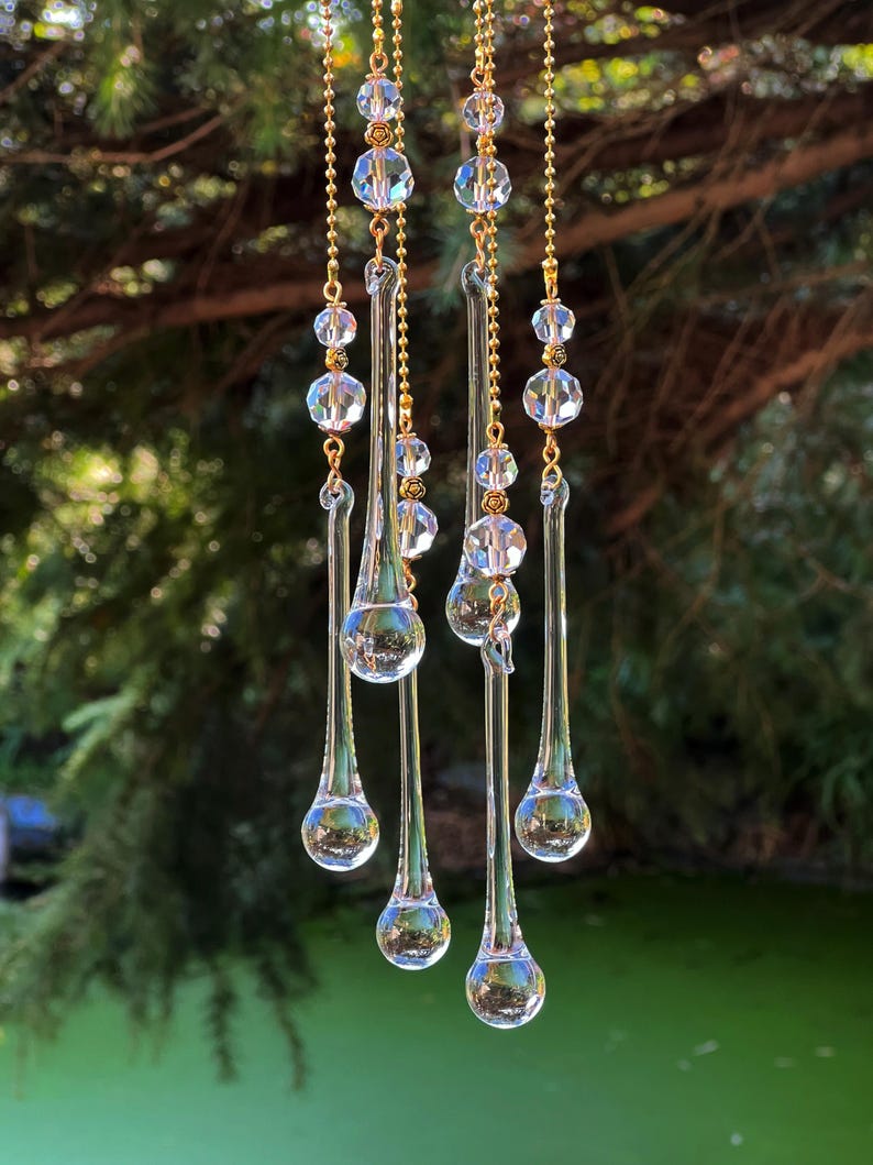 K&ouml;nnte beinhalten: Ein Kristall-Tropfen-Sonnenf&auml;nger mit f&uuml;nf Str&auml;ngen goldener Kette und klaren Kristallperlen. Der Sonnenf&auml;nger h&auml;ngt an einem Baumzweig und der Hintergrund ist ein gr&uuml;ner Teich.