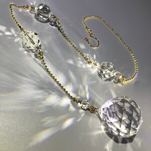 May include: A clear crystal suncatcher with a faceted sphere at the bottom and various crystal shapes along a gold-colored chain. The chain ends with a hook for hanging. The suncatcher is designed to catch and refract light.