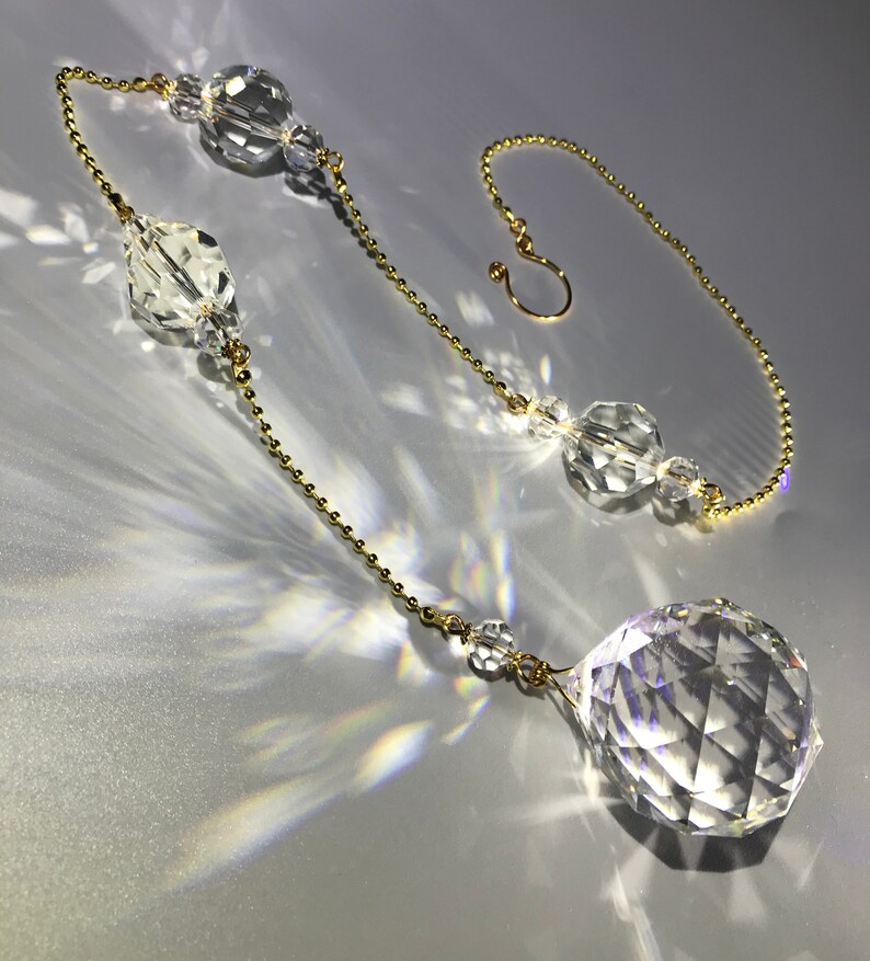 May include: A clear crystal suncatcher with a faceted sphere at the bottom and various crystal shapes along a gold-colored chain. The chain ends with a hook for hanging. The suncatcher is designed to catch and refract light.