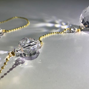 May include: Close-up of crystal prism ornaments with a gold-colored chain. The prisms are faceted, reflecting light. The chain is made of small, round beads. The background is a soft, white surface.