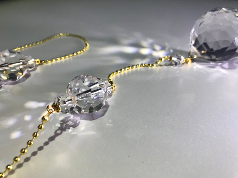 May include: Close-up of crystal prism ornaments with a gold-colored chain. The prisms are faceted, reflecting light. The chain is made of small, round beads. The background is a soft, white surface.