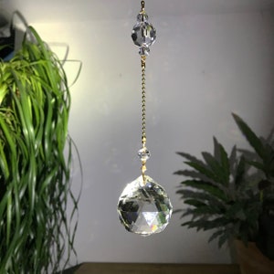 May include: A clear crystal prism suncatcher with a faceted, teardrop shape. It hangs from a gold-colored chain and is suspended between two smaller crystal spheres. The suncatcher is designed to catch and refract light, creating rainbows.