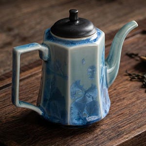 Könnte beinhalten: Eine hexagonale Keramik-Teekanne mit einer glänzenden hellblauen Glasur und dunkelblauen Kristallmustern. Die Teekanne hat einen mattschwarzen Deckel mit einem kleinen Knauf, einen geschwungenen Ausguss und einen rechteckigen Henkel.