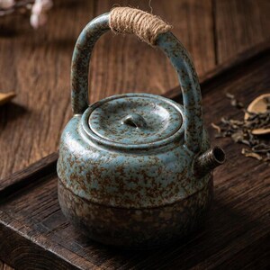 May include: A ceramic teapot with a speckled blue and brown glaze, featuring a rounded body, a matching lid, and a high arched handle wrapped in natural jute twine. The teapot has a short, straight spout and a textured, rustic finish.