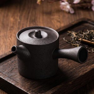 Può includere: Una teiera in ceramica marrone scuro dalla superficie ruvida, con corpo cilindrico, coperchio piatto con un piccolo pomello centrale, beccuccio corto e un lungo manico laterale dritto che si estende orizzontalmente, finitura opaca.