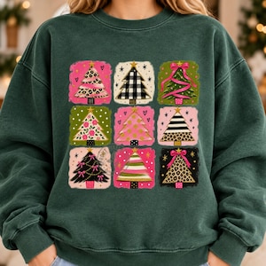 May include: A dark green crewneck sweatshirt featuring a graphic print of nine distinct Christmas tree designs arranged in a grid. Each tree is set against a square background with varied patterns including leopard print, plaid, floral, and stripes.