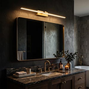 May include: A rectangular wall mirror with a thin gold frame mounted above a dark marble vanity countertop. A long, linear gold LED light fixture is installed horizontally above the mirror, casting a warm glow over the bathroom area.