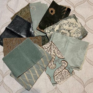 May include: A collection of fabric swatches featuring various textures and patterns, including sage green, dark charcoal, brown, and cream tones. The pieces display diverse designs such as floral motifs, geometric grids, and abstract shapes.