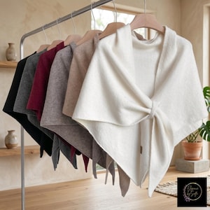 May include: A row of soft, triangular shawls hanging on a metal rack, featuring a variety of colors including cream, beige, gray, burgundy, dark gray, and black. Each shawl has a knotted front detail and a small tag near the bottom edge.