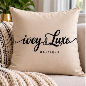 May include: A square beige throw pillow featuring the black text "ivey & Luxe" in a stylized script font, with the word "Boutique" printed in a smaller, simple sans-serif font directly underneath the main text.