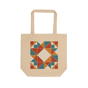 May include: A beige tote bag featuring a centered geometric pattern composed of triangles and squares in shades of burnt orange, teal, and mustard yellow. The design creates a symmetrical, quilt-like motif with a large diamond-shaped negative space.