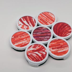 May include: A cluster of seven circular phone grips arranged in a flower-like pattern. Each grip features a white plastic rim and a flat top surface decorated with abstract, textured brushstroke patterns in various shades of red, orange, and white.