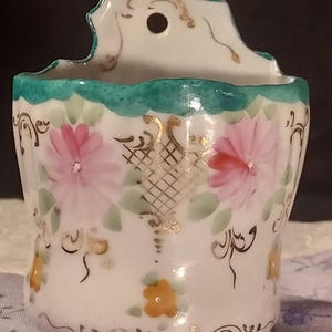 May include: A white ceramic wall pocket featuring a scalloped teal rim and a central hole for hanging. The exterior is decorated with hand-painted pink flowers, small yellow blossoms, green leaves, and gold filigree accents in a lattice pattern.