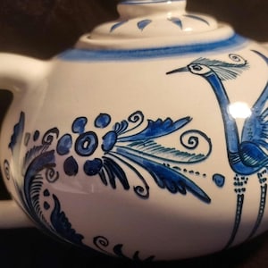 May include: A white ceramic teapot featuring hand-painted blue floral patterns and a stylized blue bird design. The teapot includes a matching lid with a blue rim, a curved handle, and a spout, all decorated with intricate blue brushwork.