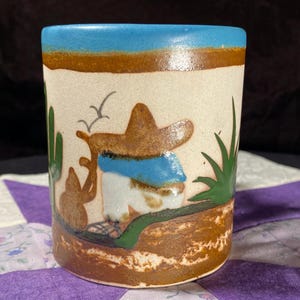 Pode incluir: Uma caneca de cerâmica com uma cena de deserto pintada à mão, apresentando uma figura com um chapéu sombrero castanho, cactos verdes e uma faixa de céu azul. A base e a borda são acentuadas com tons de terra castanhos e um edifício.