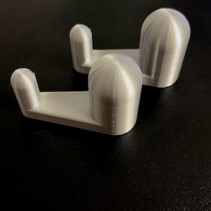 Peut inclure: Deux crochets argentés en forme de L avec un sommet arrondi et une base plate. Chaque crochet présente un montant vertical relié à un bras horizontal, avec une texture imprimée en 3D visible sur la surface du matériau plastique.