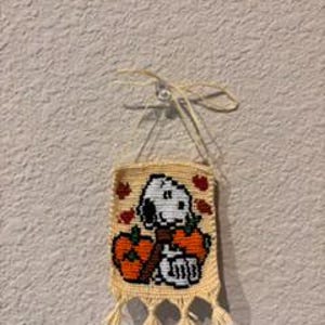 以下が含まれることがあります： クリーム色のクロッシェ編みの壁掛け。黒い耳の白い犬が2つのオレンジ色のカボチャの間に座っているクロスステッチのデザインが施されています。小さな赤い葉のアクセント、上部の吊り下げ用紐、4つのタッセルが付いています。