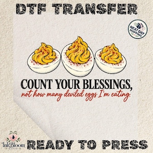 May include: A DTF transfer sheet featuring three deviled eggs with yellow yolks and red paprika seasoning. The text reads: DTF TRANSFER, COUNT YOUR BLESSINGS, not how many deviled eggs I'm eating, NEXT DAY SHIPPING, and READY TO PRESS.