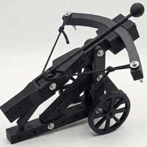 Pode incluir: Um modelo de balista em miniatura preto com rodas, construído com uma estrutura, um mecanismo de disparo central e uma corda de tensão. O dispositivo possui uma roda de raios em cada lado, parafusos metálicos prateados nas juntas e um projétil arredondado.