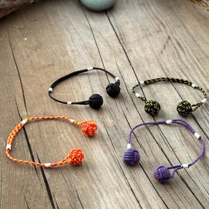 May include: Four bracelets made of cord, each featuring two spherical monkey fist knots at the ends. The bracelets come in solid black, yellow and black patterned, red and yellow patterned, and blue and red patterned cord with small white accents.