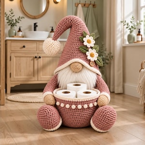 May include: A crocheted gnome figure in dusty rose with a cream-colored pom-pom on its tall hat, adorned with two white daisy flowers and green leaves. The gnome has a beige nose, a white beard, and holds a matching basket containing three rolls of toilet paper.