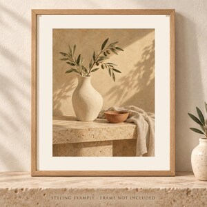 May include: A framed wall art print featuring a textured off-white vase holding olive branches with green leaves and small olives. Next to the vase sits a small terracotta-colored bowl and a draped beige fabric cloth on a stone surface.