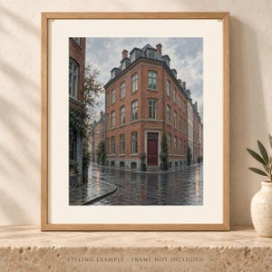 May include: A framed wall art print depicting a multi-story red brick building on a street corner. The structure features numerous windows, a dark red front door, and a gray roof. The wet cobblestone street reflects the overcast sky and building.