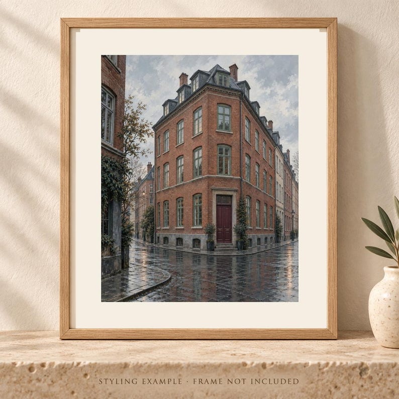 May include: A framed wall art print depicting a multi-story red brick building on a street corner. The structure features numerous windows, a dark red front door, and a gray roof. The wet cobblestone street reflects the overcast sky and building.