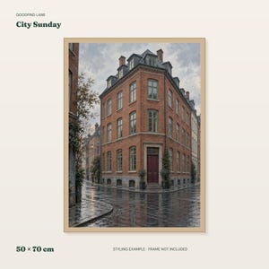 May include: A framed art print titled City Sunday by Goodfind Lane, measuring 50 x 70 cm. The image depicts a multi-story red brick corner building with a dark red door and grey roof, situated on a wet, reflective cobblestone street under a cloudy sky.