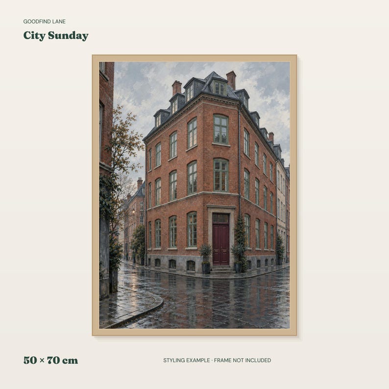 May include: A framed art print titled City Sunday by Goodfind Lane, measuring 50 x 70 cm. The image depicts a multi-story red brick corner building with a dark red door and grey roof, situated on a wet, reflective cobblestone street under a cloudy sky.
