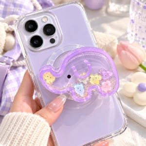May include: A clear smartphone case with a light purple back panel featuring a purple elephant-shaped phone grip. The grip is filled with liquid and small, colorful floating cloud-shaped charms in yellow, blue, pink, and white, creating a shaker effect.