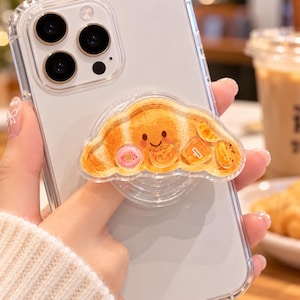 May include: A clear smartphone case featuring a collapsible phone grip shaped like a golden-brown croissant with a smiling face. The croissant graphic includes small illustrations of a pink donut, a bagel, a jar of honey, and a slice of orange.