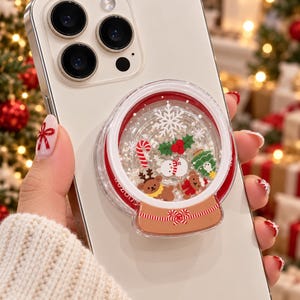May include: A white smartphone featuring a circular, clear liquid-filled phone grip shaped like a snow globe. Inside the globe are miniature floating figures including a candy cane, a snowman, a green tree, a reindeer, and a bear, with a snowflake.
